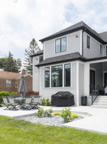 The backyard of a beautiful grey, luxury house with black accents, a covered seating area, large concrete patio, and landscaping